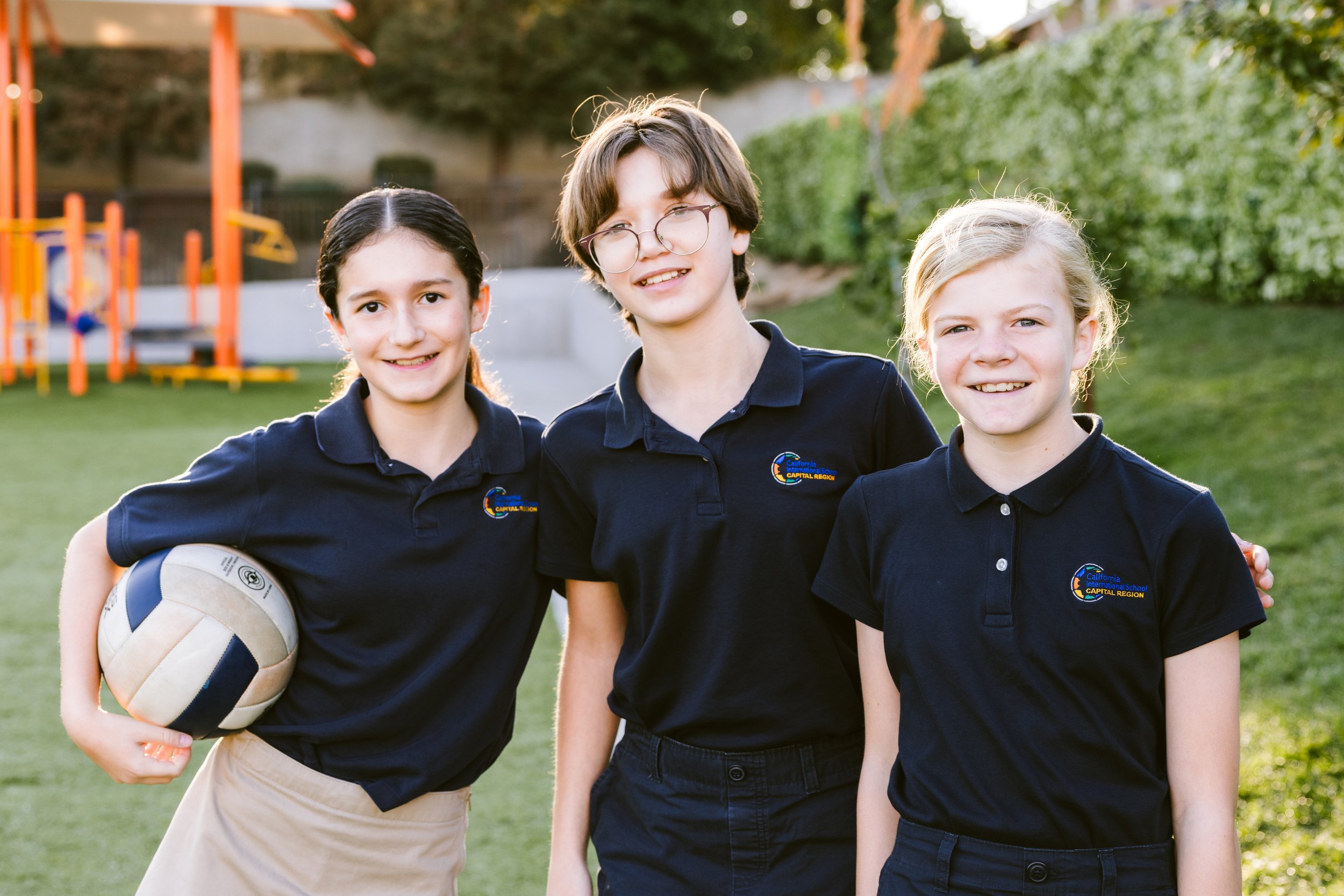 Three smiling CIS students in school uniforms posing outdoors with playground equipment visible in background